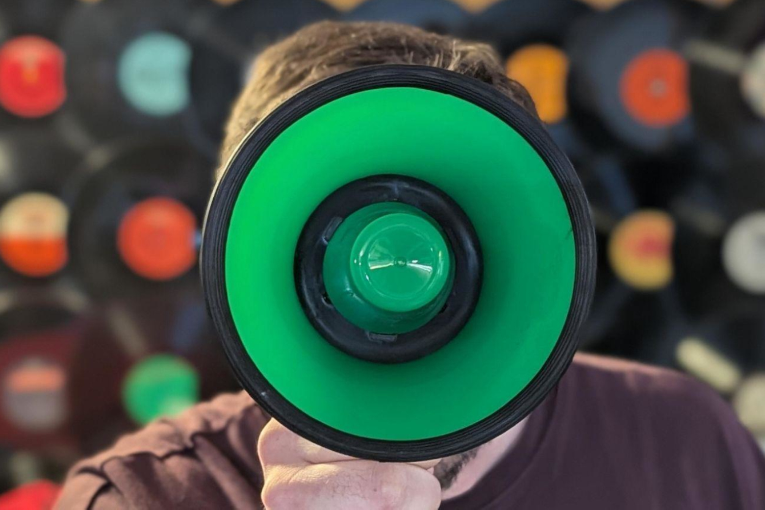 Vocalist with megaphone in front of a wall full of records. His head is hidden behind the megaphone.